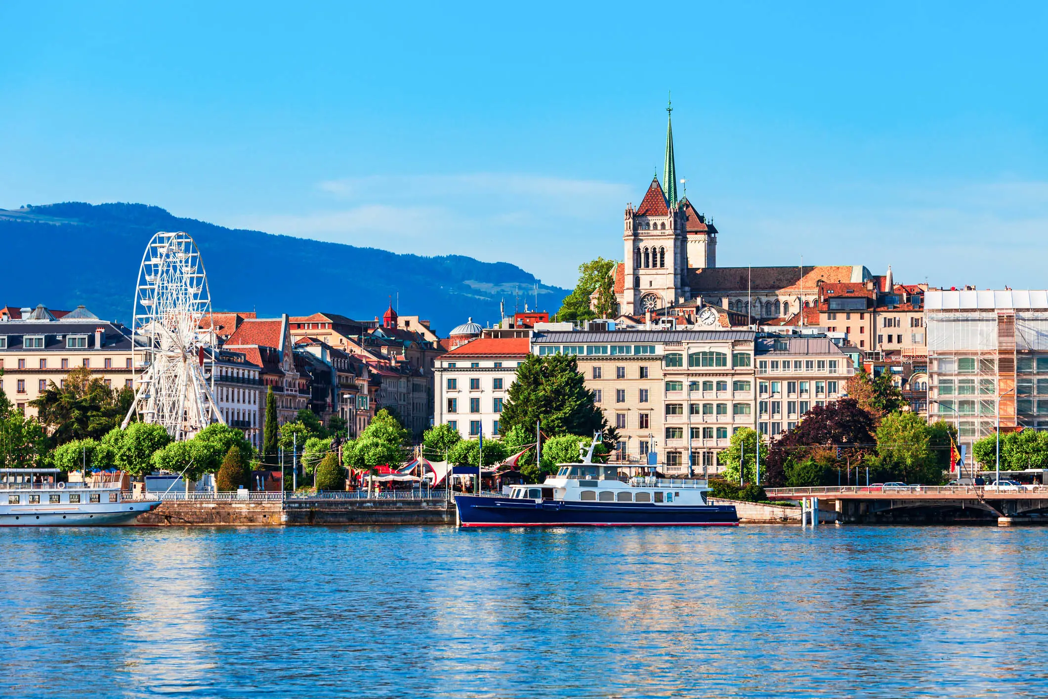 Aerial view, Geneva, Switzerland Geneva cityscape with cathedral spire, ferris wheel, and waterfront buildings along Lake Geneva under blue sky.