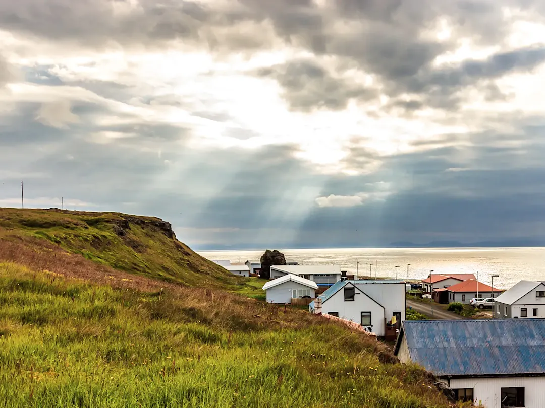 Küstenlandschaft mit Häusern und Sonnenstrahlen durch die Wolken. Skagaströnd, Norðurland vestra, Island.