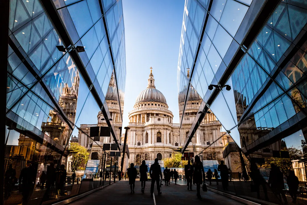 Menschen gehen zwischen zwei modernen Glasgebäuden, in denen sich die ikonische Kuppel der St. Paul's Cathedral in London spiegelt, unter einem klaren blauen Himmel. Die Perspektive führt den Blick auf die großartige Architektur der Kathedrale.