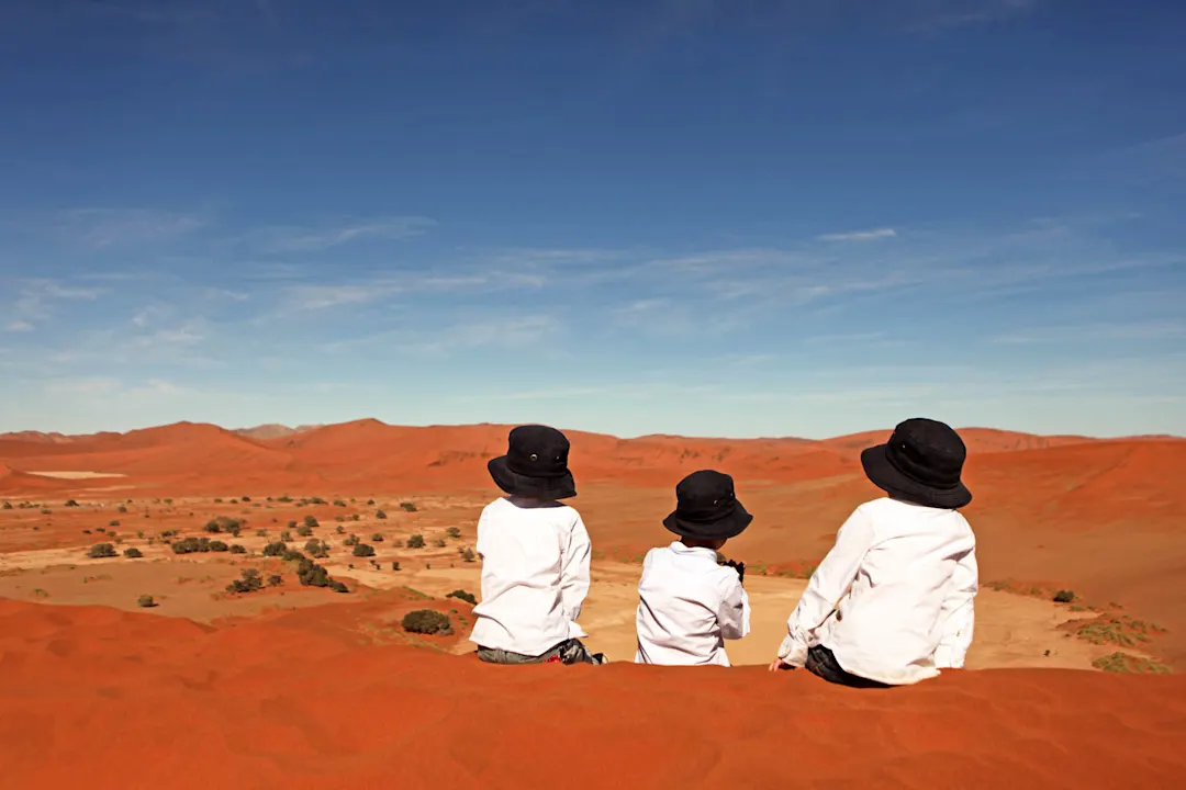 Drei Personen mit schwarzen Hüten und weißen Hemden sitzen auf einer roten Sanddüne und betrachten die Wüstenlandschaft.