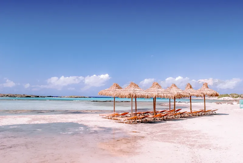 Sandy beach with thatched umbrellas and sun loungers in front of turquoise waters. Elafonisi beach, Greece.