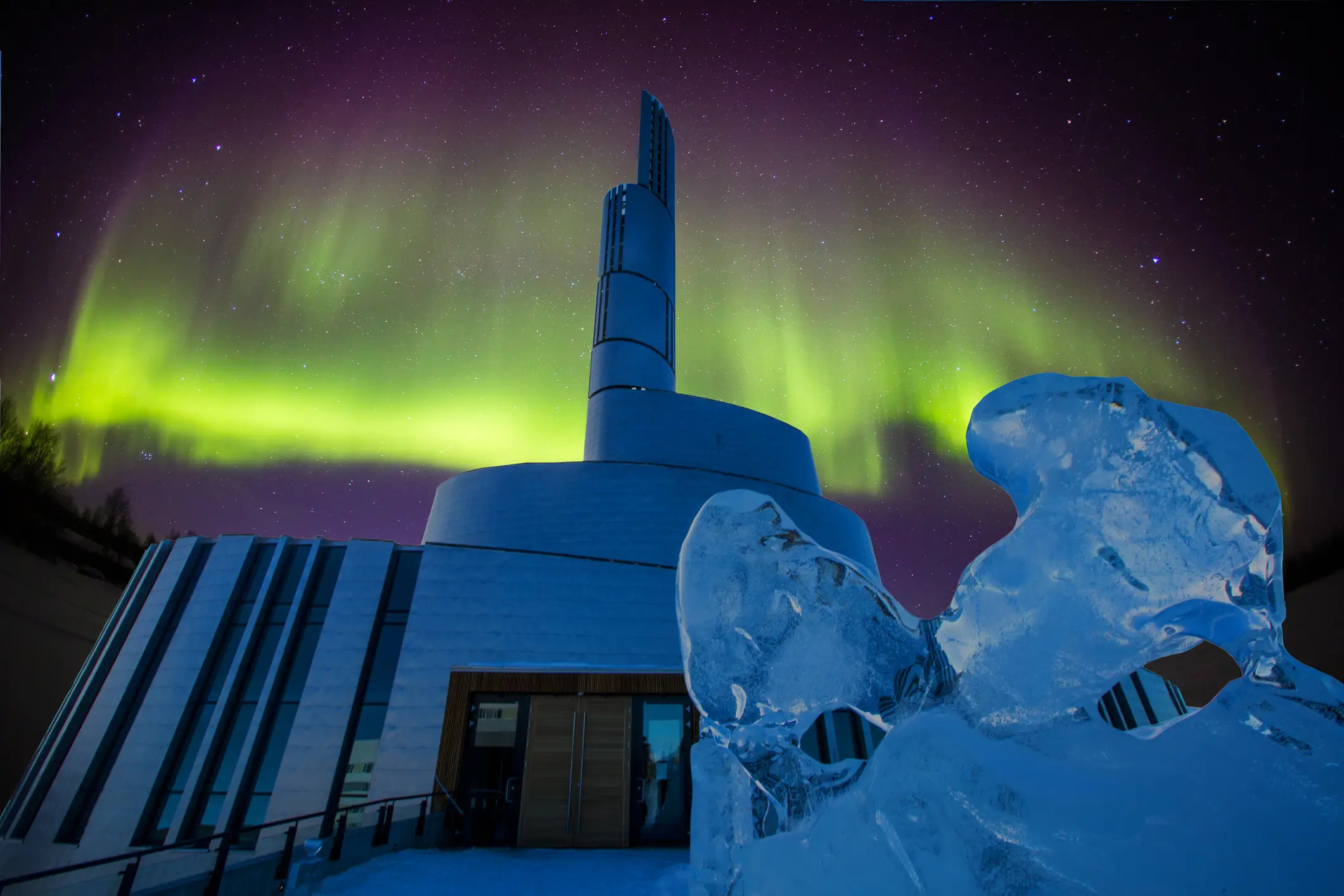 Eine moderne weiße Kirche mit rundem Turm und im Hintergrund ein von grün-gelblich schimmernden Nordlichtern erhellter Himmel