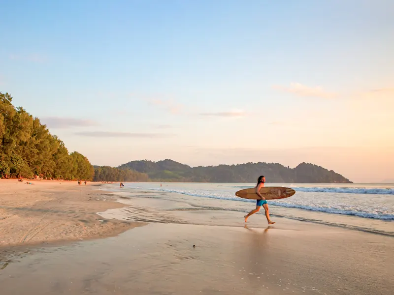 Thaïlande, Surfeur Surfeur avec planche de surf marchant le long de la plage au coucher du soleil avec de douces vagues en arrière-plan. Thaïlande.