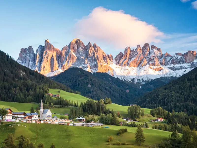 Village avec une église à haut clocher dans une vallée verdoyante surplombée par des montagnes enneigées. Dolomites, Italie.