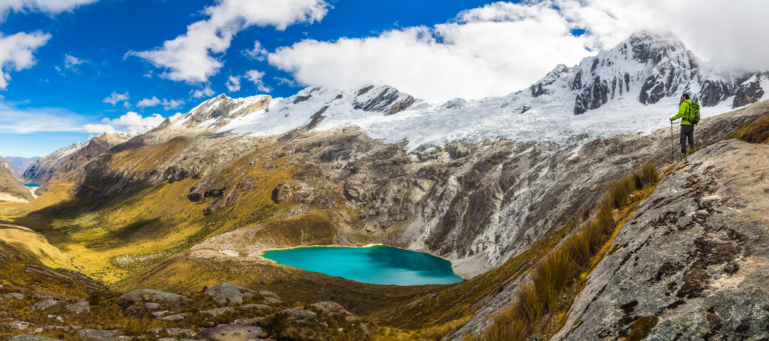Männlicher Abenteurer mit einem Rucksack auf einem Hügel auf der rechten Seite mit Blick auf die wunderschöne und weite Tallandschaft mit einem türkisblauen Aquarellsee in Cordillera Blanca, Peruanische Berge, Südamerika