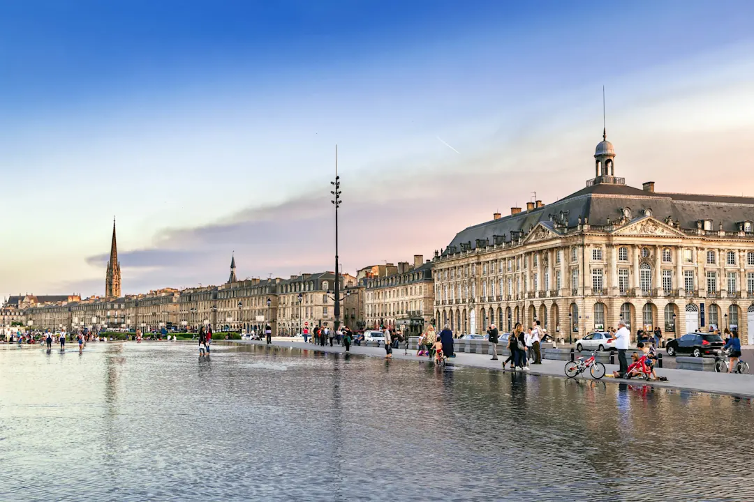 Place de la Bourse in Bordeaux mit Wasserspiegel und historischen Gebäuden bei Sonnenuntergang, Menschen spazieren am Ufer.