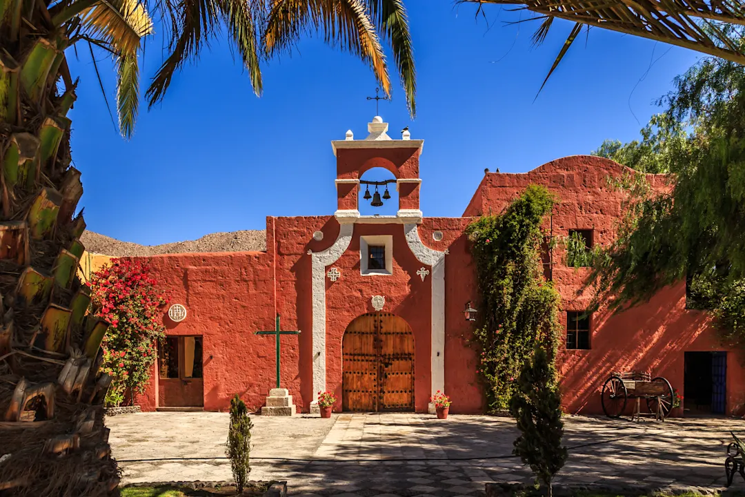 Rote Wände der spanischen katholischen Kapelle mit Palmen, Bäumen und Blumen, Arequipa, Peru

