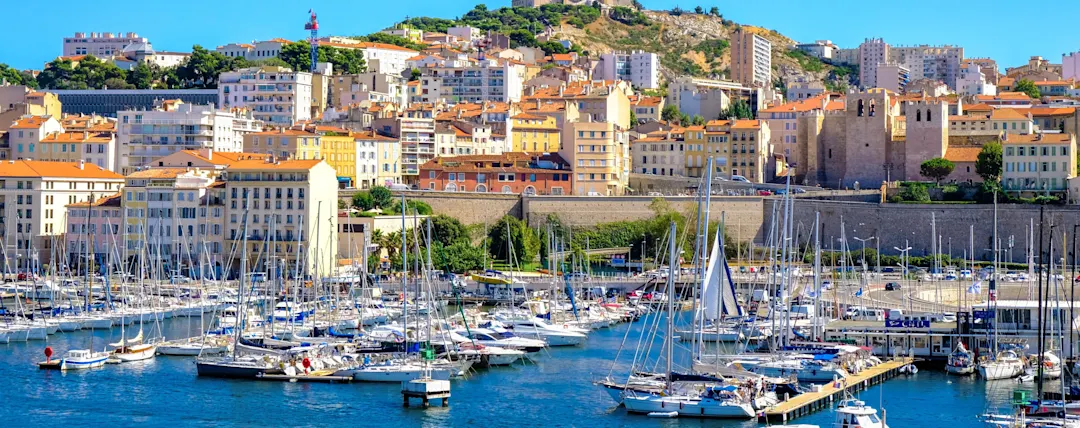 Panoramablick auf Marseille mit Hafen voller Segelboote, bunten Gebäuden und der Notre-Dame de la Garde Basilika auf dem Hügel.