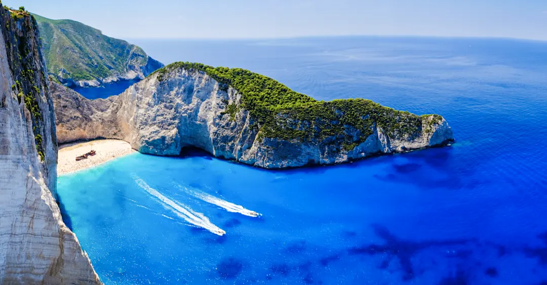 Luftaufnahme einer Klippe mit einem Strand und türkisfarbenem Wasser drumherum, mit mehreren Booten.