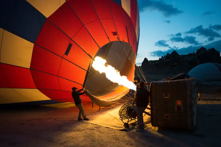 Zwei Personen blasen in der Morgendämmerung einen großen Heißluftballon mit einer Flamme auf. Der Ballon leuchtet orange und rot gegen den frühen Morgenhimmel, während ein Weidenkorb in der Nähe auf der Seite ruht. Im Hintergrund ist felsiges Gelände zu sehen.