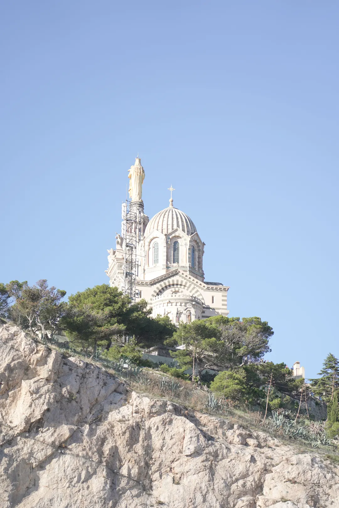 Weiße Kathedrale Notre-Dame de la Garde auf felsigem Hügel mit Kuppel und Marienstatue unter blauem Himmel.