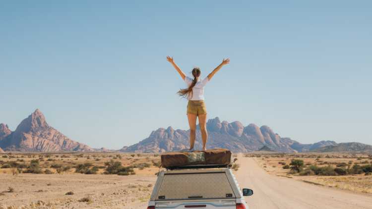Frau streckt die Arme in die Luft auf dem Dach eines Autos in der namibischen Landschaft während eines Roadtrips