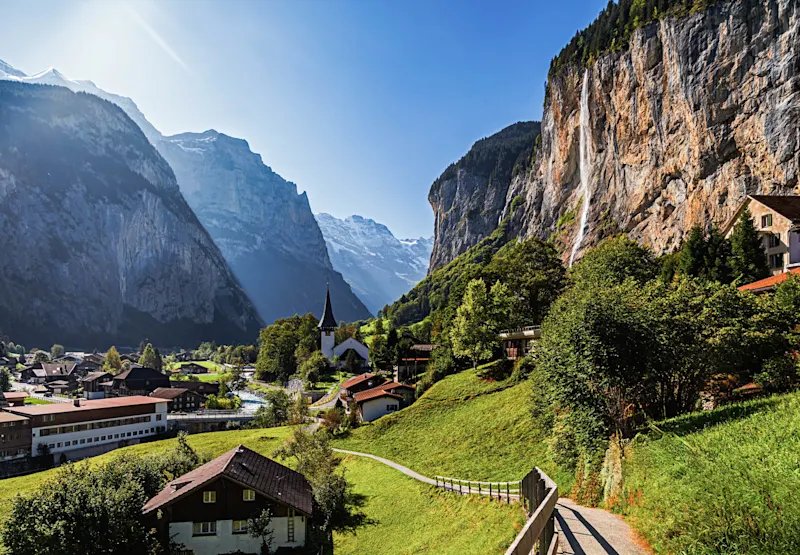 Swiss alpine village nestled in Lauterbrunnen Valley with towering cliffs, waterfall, church steeple, and snow-capped mountains.