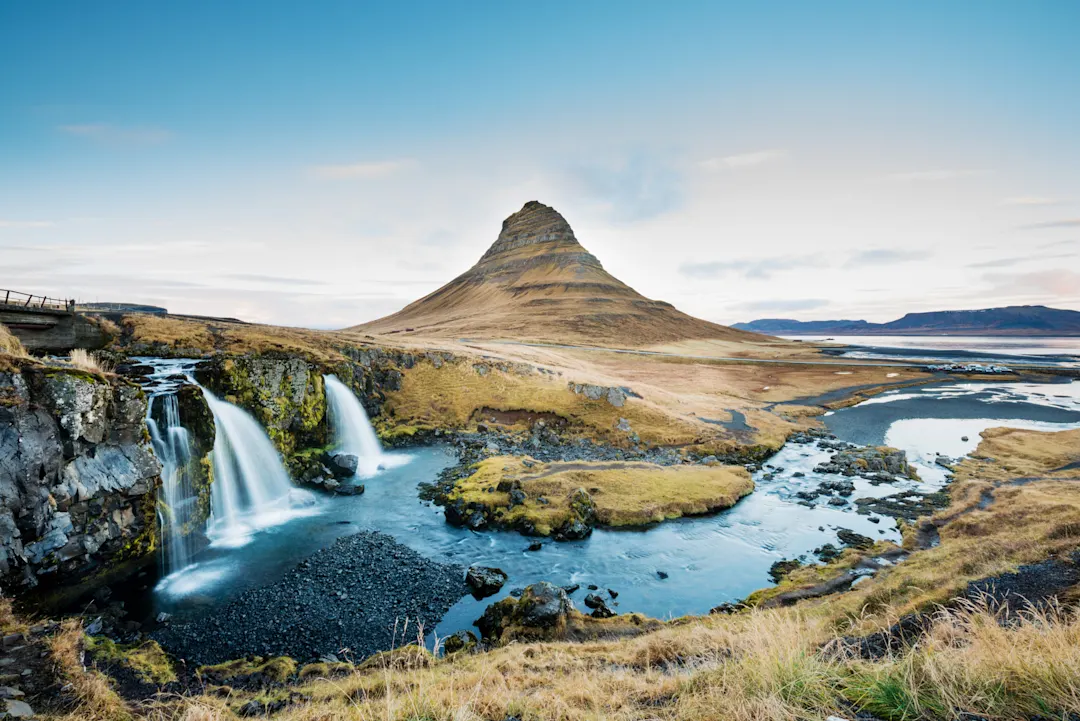 Der Berg Kirkjufell in Island mit Wasserfällen im Vordergrund unter blauem Himmel und weiter Landschaft.