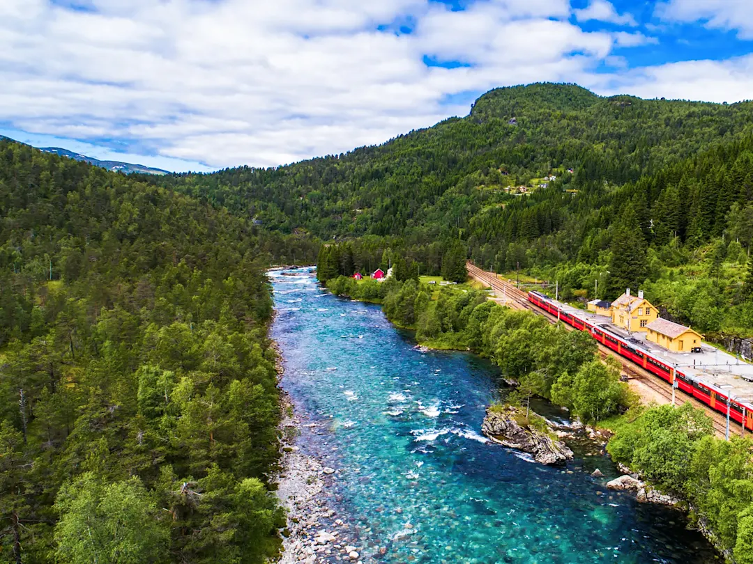 Roter Zug an malerischem Fluss in grüner Landschaft. Bjorli, Innlandet, Norwegen.