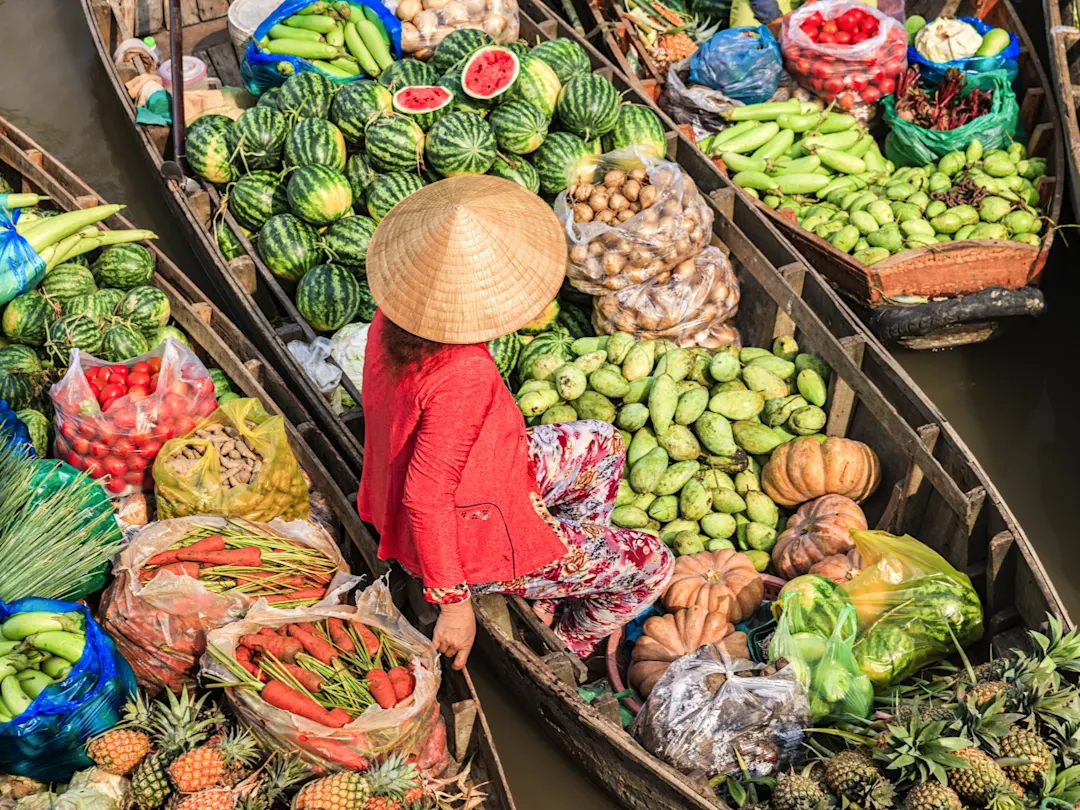 Frau mit Non-La-Hut auf einem schwimmenden Markt, umgeben von frischem Obst und Gemüse. Mekong-Delta, Vietnam.