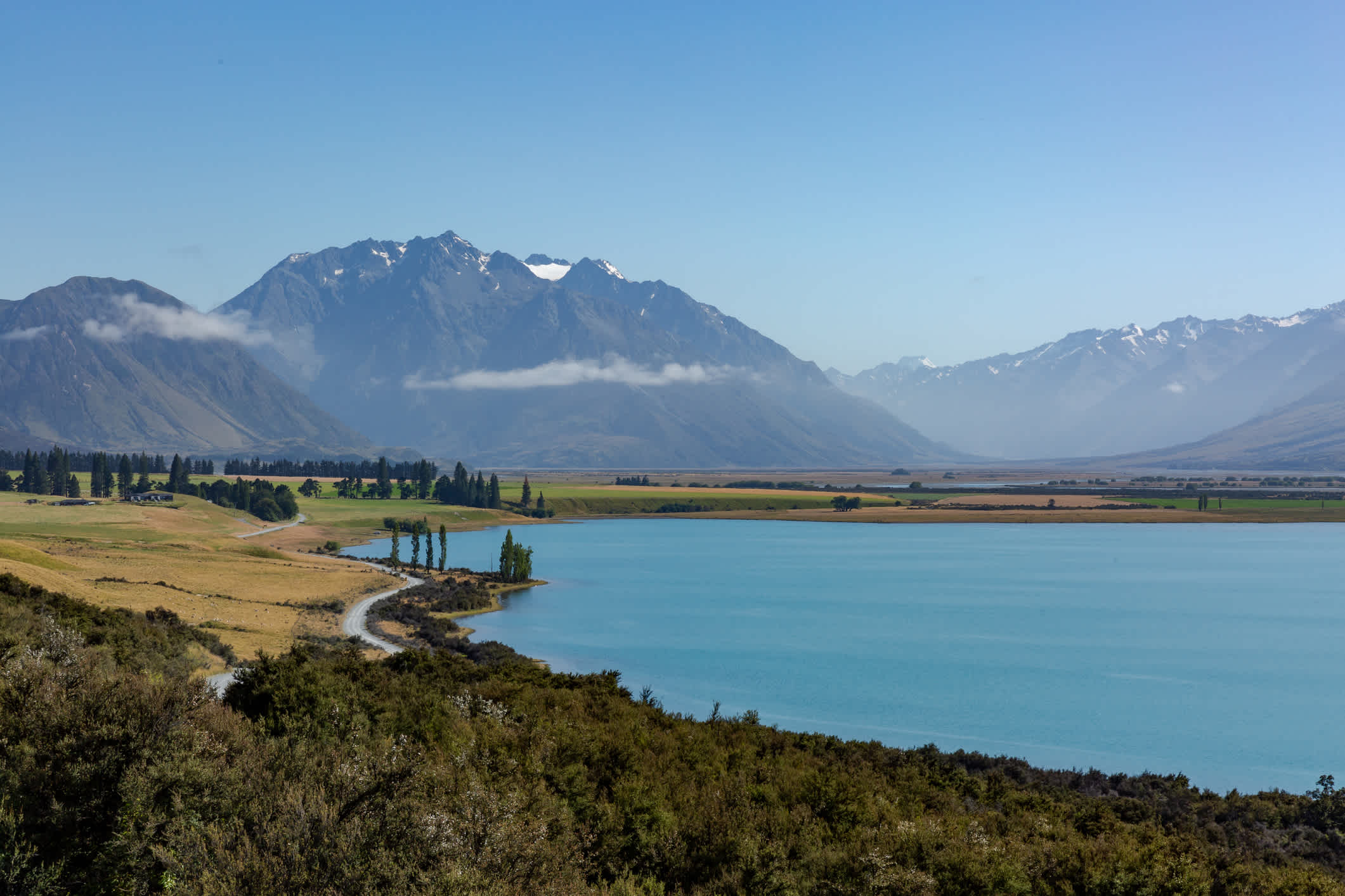 Ein Foto des türkisfarbenen Wassers des Ohau-Sees mit den Südalpen im Hintergrund. Kurvenreiche Straße entlang des Sees, morgens tiefhängende Wolken im Tal.