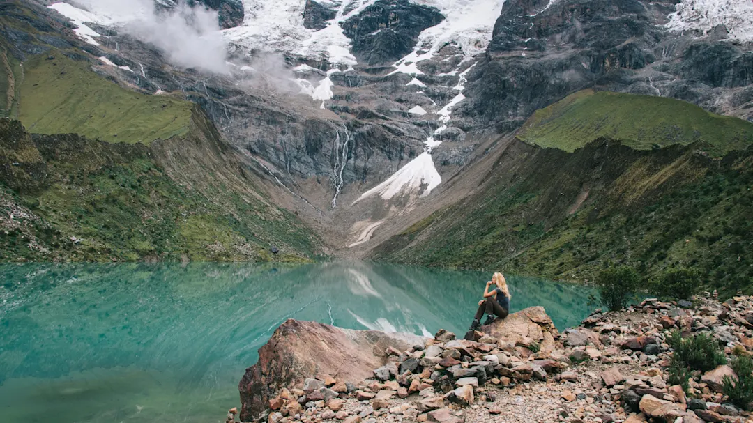 Frau sitzt an einem türkisfarbenen Bergsee mit schneebedeckten Gipfeln. Salcantay, Cusco, Peru.