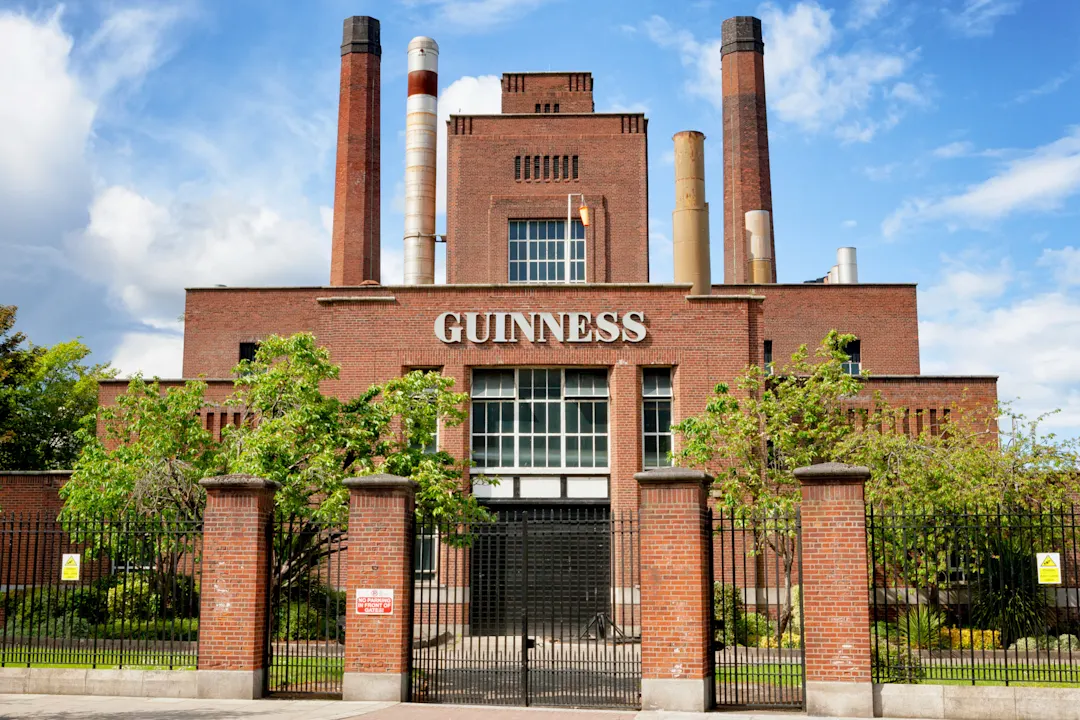 Guinness Storehouse Das Bild zeigt das Äußere der Guinness-Brauerei in Dublin, Irland, mit dem roten Backsteingebäude, den hohen Schornsteinen, dem Eingangstor und den grünen Bäumen davor unter einem teilweise bewölkten Himmel.