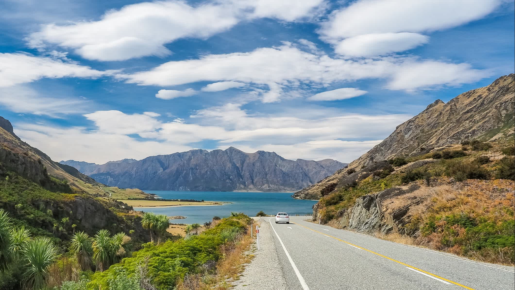 Voiture sur la route de The Neck Lookout bordée de montagnes avec vue du lac Hawea en arrière-plan, Nouvelle-Zélande