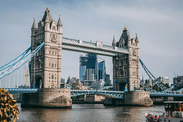 Das Bild zeigt Londons ikonische Tower Bridge, die die Themse überspannt, mit modernen Wolkenkratzern im Hintergrund unter einem bewölkten Himmel. Ein kleines Boot ist teilweise auf der unteren rechten Seite des Flusses zu sehen.