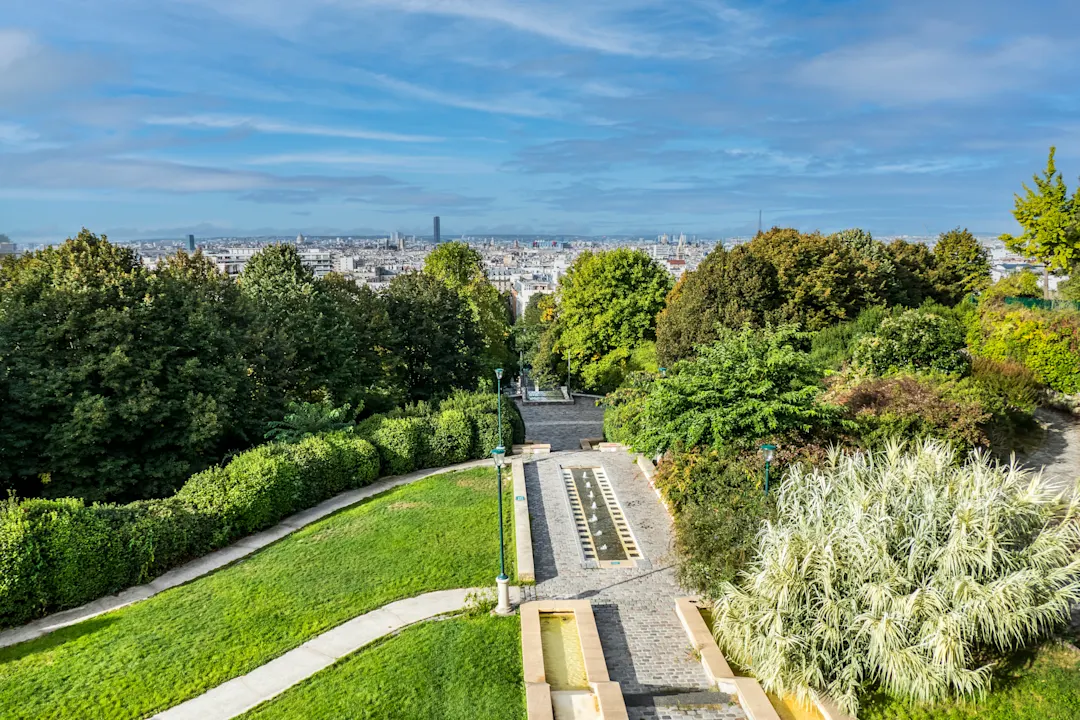 Panoramablick auf Paris von einem grünen Park mit gepflegten Wegen, Brunnen und üppiger Vegetation bei blauem Himmel.
