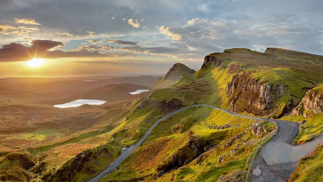 Ein malerischer Blick auf eine kurvenreiche Straße durch grüne Hügel bei Sonnenaufgang, mit felsigen Klippen, kleinen Seen und einem dramatischen Himmel voller Wolken und Sonnenlicht.