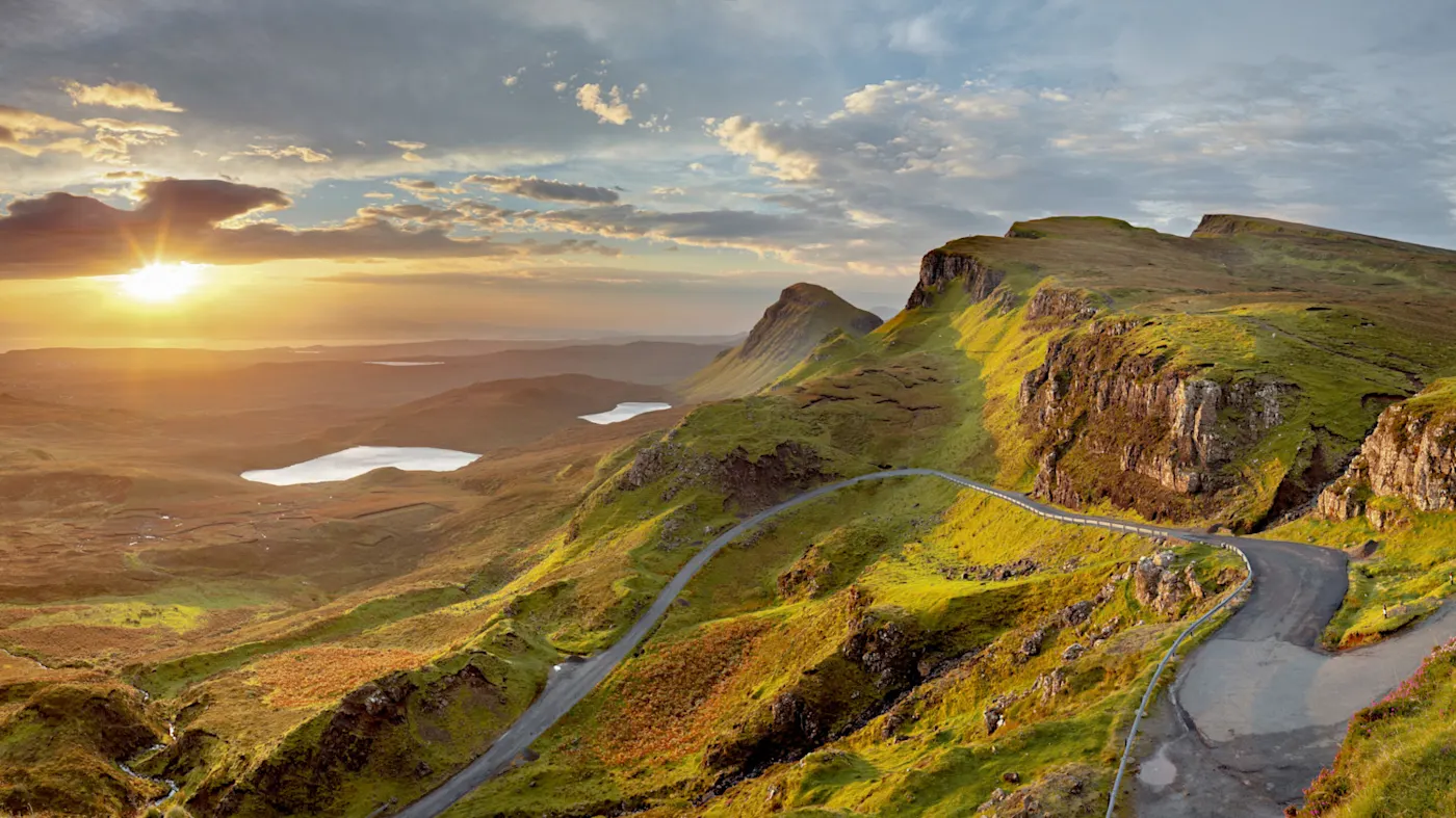 isle of skye Ein malerischer Blick auf eine kurvenreiche Straße durch grüne Hügel bei Sonnenaufgang, mit felsigen Klippen, kleinen Seen und einem dramatischen Himmel voller Wolken und Sonnenlicht.