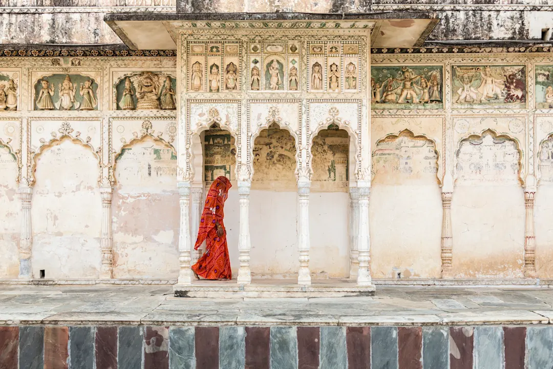 Person in orangerotem Sari vor historischer indischer Palastfassade mit kunstvollen Wandmalereien und Bogengängen.