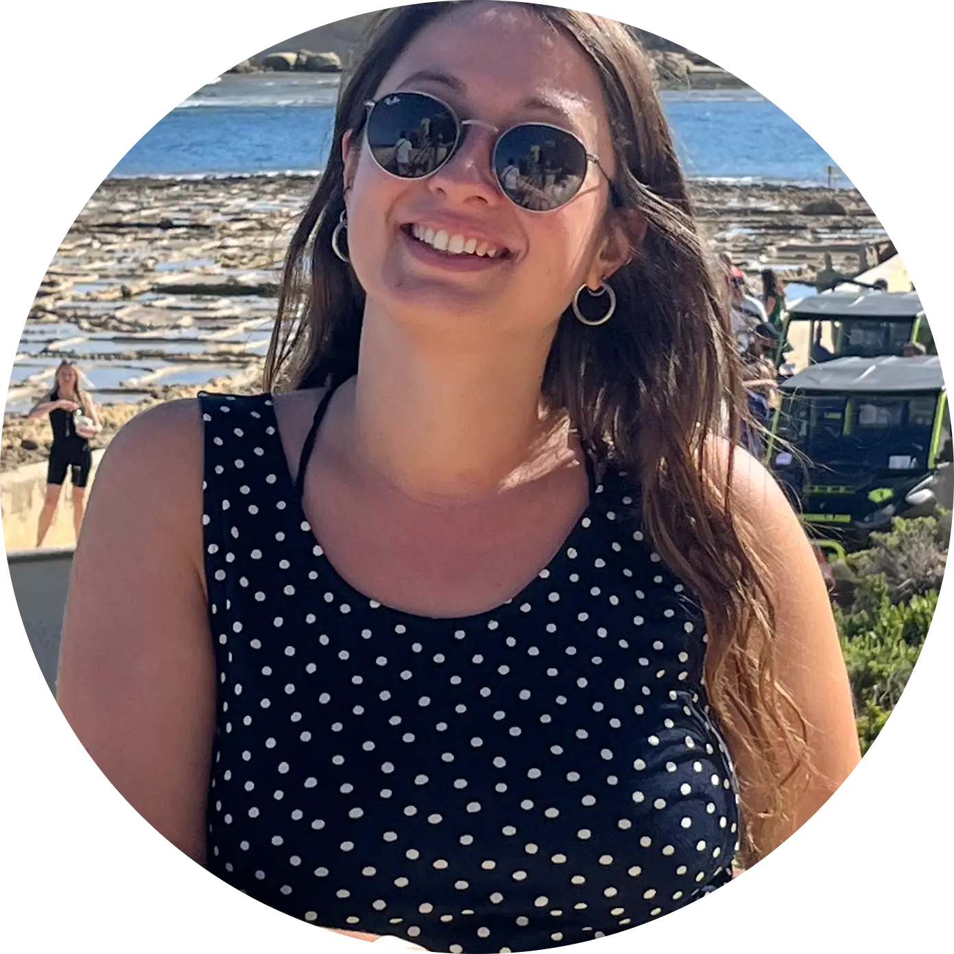 Lisa Robin, in polka dot dress and sunglasses at coastal viewpoint with rocky shoreline and distinctive flat-top formation in background.