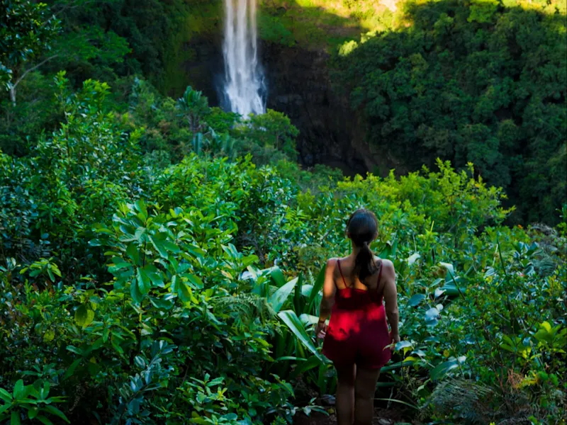 Frau in rotem Kleid blickt auf hohen Wasserfall in tropischer Landschaft, Chamarel, Mauritius