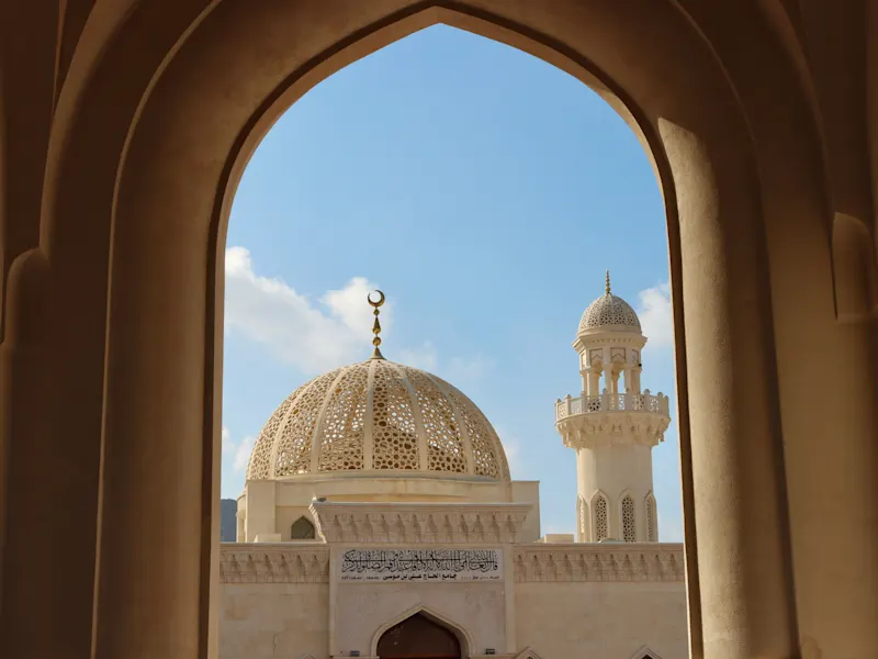 Blick durch einen Bogen auf eine Moschee mit verzierter Kuppel und Minarett, Maskat, Oman