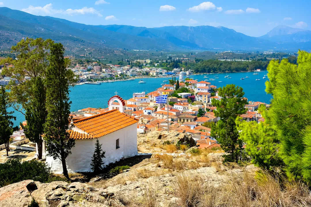 Panoramablick auf eine griechische Küstenstadt mit weißen Häusern, türkisblauem Meer und Bergen im Hintergrund.