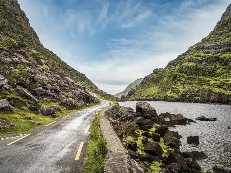 Serpentine road through mountainous terrain with water and rocks on the banks. Kerry, Ireland.