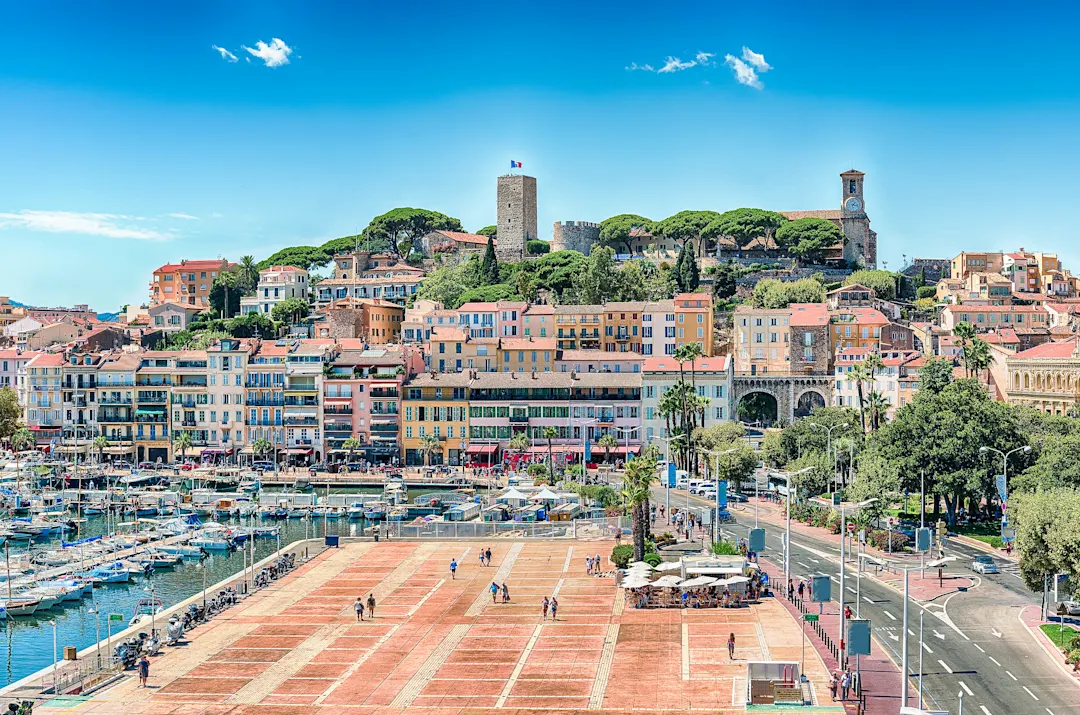Harbor, Cannes, Cote Azur Panoramablick auf Cannes mit bunten Häusern am Hügel, historischem Turm, Yachthafen und Promenade unter blauem Himmel.