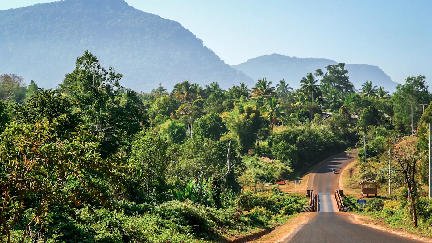 Bolaven Plateau, Laos Asphaltstraße durch üppig grüne Vegetation mit Bergen im Hintergrund, Bolaven Plateau, Laos.