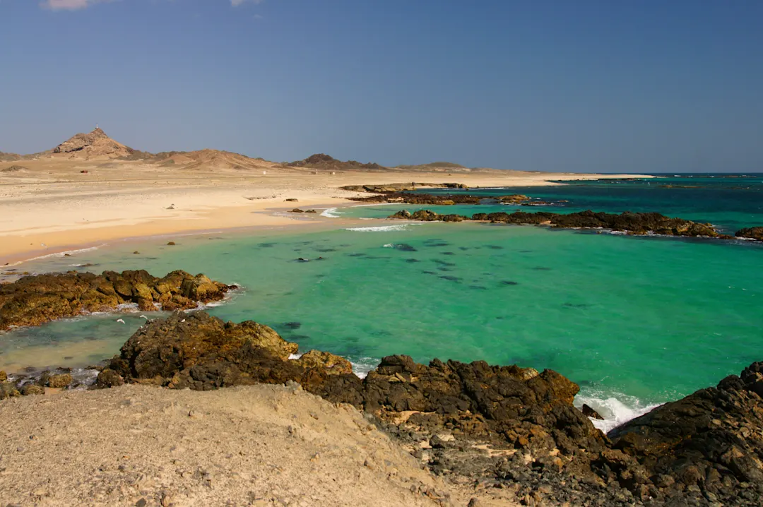 Karge Küstenlandschaft mit türkisfarbenem Wasser und Felsen, die den goldenen Sandstrand umgeben, Masirah Island, Oman.