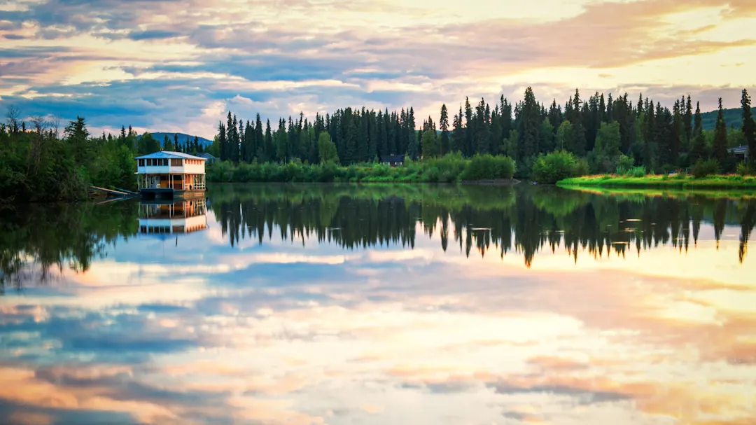 Spiegelung eines Hauses und der umgebenden Bäume bei Sonnenuntergang in einem See. Fairbanks, Alaska, USA.