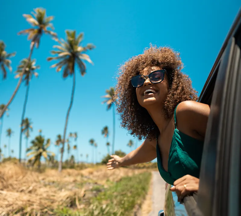 Reiseart, Roadtrip Eine Frau lächelt und schaut aus dem Auto. Im Hintergrund sind Palmen.