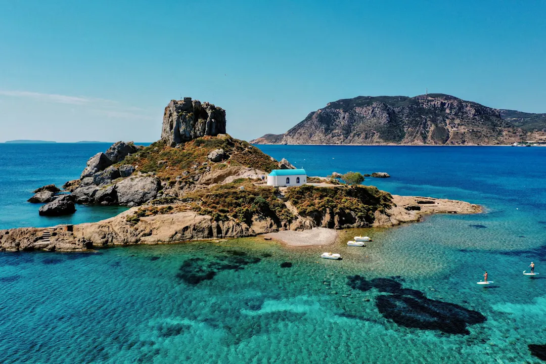 Kleine Insel mit weißer Kapelle und Felsformation im türkisblauen Meer vor griechischer Küste, einige Paddler im Wasser.