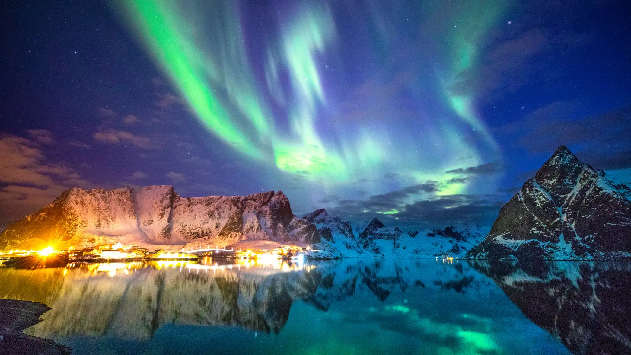 Grünes Polarlicht am Himmel über einem Fischerdorf an einem Fjord mit schneebedeckten Bergen im Hintergrund