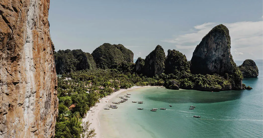 Ein tropischer Strand mit klarem türkisfarbenem Wasser, weißem Sand und Booten, die in Ufernähe vor Anker liegen. Im Hintergrund erheben sich üppig grüne Klippen und felsige Kalksteinformationen unter einem sonnigen, klaren Himmel.