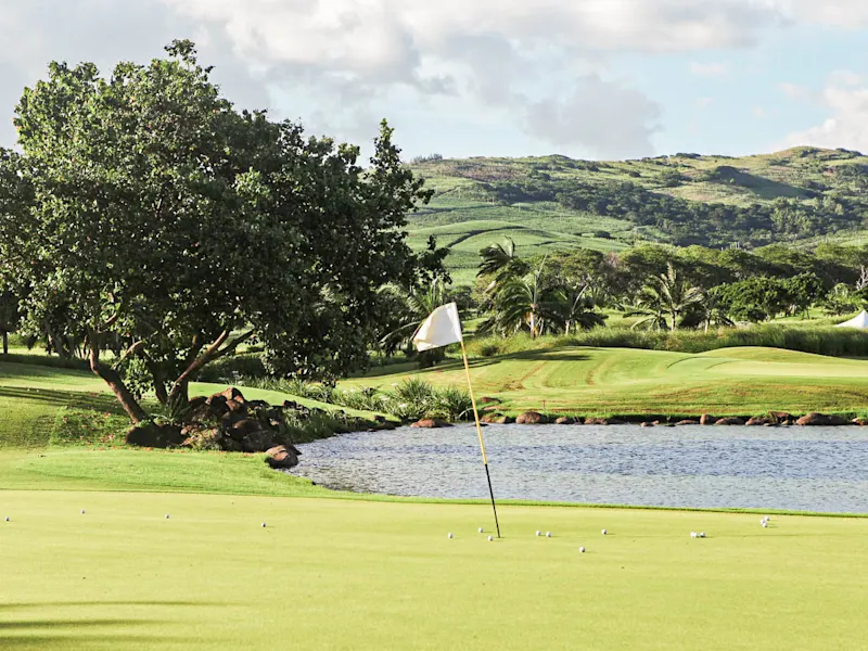 Terrain de golf avec pelouse verte, lac et collines en arrière-plan, entouré de végétation tropicale. Île Maurice.