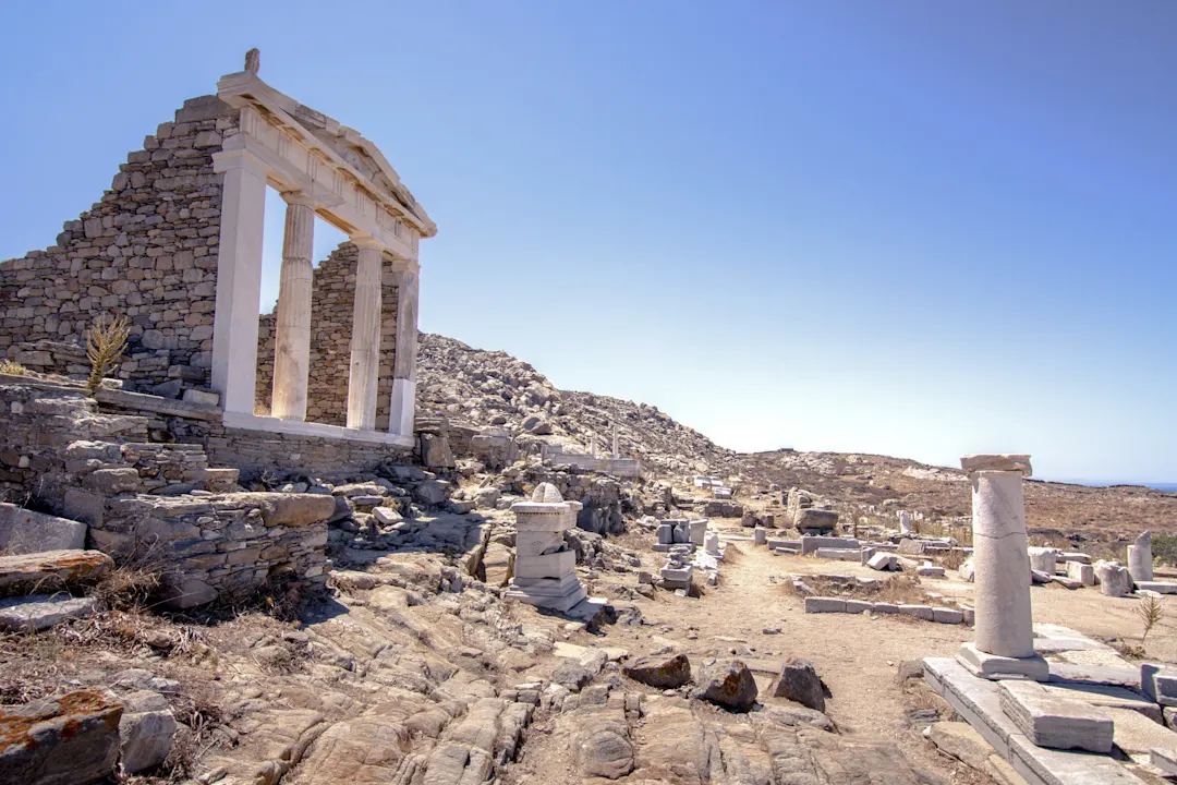 Delos Alte Steinruinen mit weißen Säulen unter einem klaren blauen Himmel, verstreut über eine felsige, sonnenbeschienene Landschaft mit Resten von Säulen und Mauern im Hintergrund.