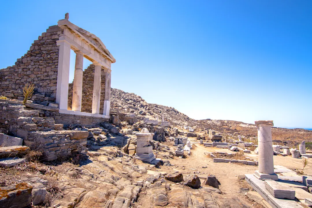 Delos Alte Steinruinen mit weißen Säulen unter einem klaren blauen Himmel, verstreut über eine felsige, sonnenbeschienene Landschaft mit Resten von Säulen und Mauern im Hintergrund.