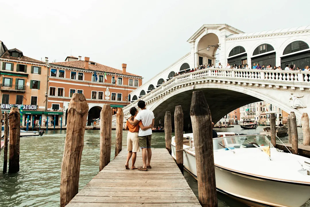 Paar betrachtet die Rialtobrücke in Venedig von einem Holzsteg aus, umgeben von Kanälen und historischen Gebäuden.