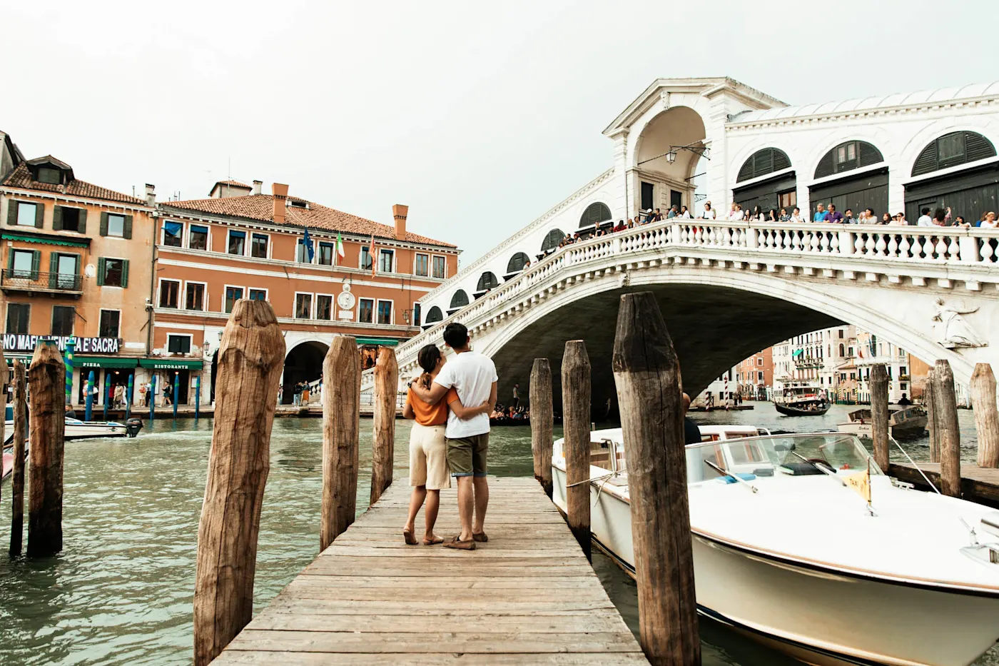 Couple admirant le pont du Rialto à Venise depuis un ponton en bois, avec des bateaux amarrés et des bâtiments colorés.