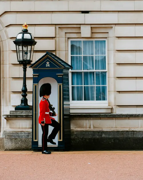 Eine britische königliche Wache in roter Uniform und Bärenfellmütze steht stramm vor einem Wachhäuschen in der Nähe eines Fensters und einer schwarzen Lampe im Buckingham Palace.