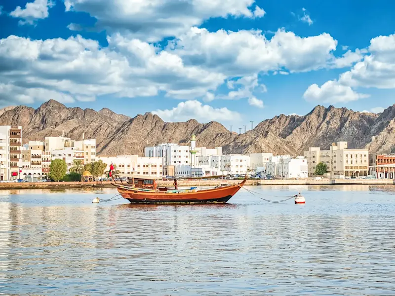 Traditionelles Boot vor modernen Gebäuden und Bergen. Muscat, Muscat Governorate, Oman.
