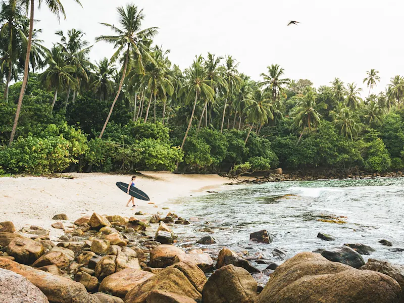 Une surfeuse avec une planche de surf noire se promène le long d'une plage de sable isolée avec des palmiers au Sri Lanka.
